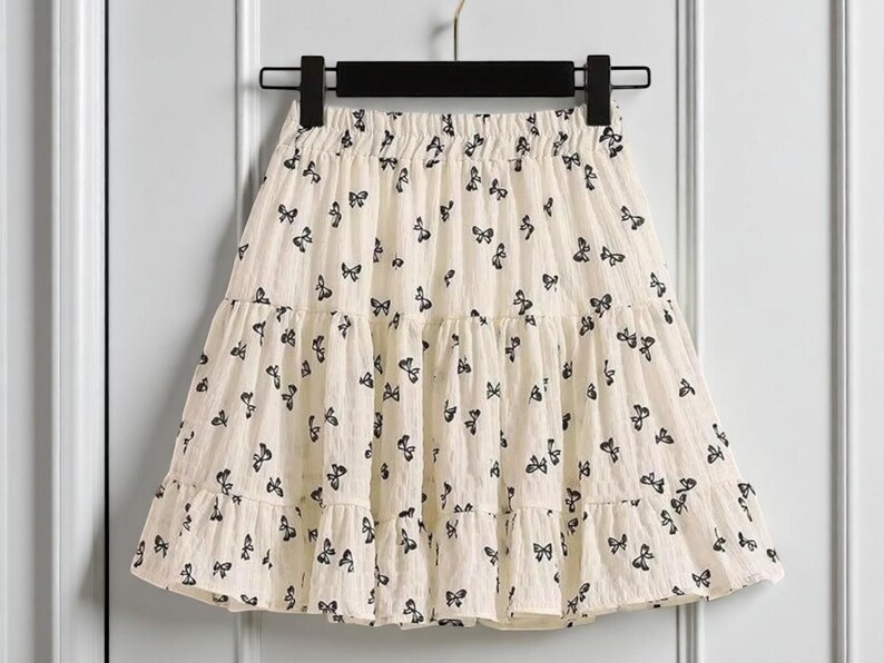 May include: A cream-colored skirt with a tiered design, adorned with a repeating pattern of black butterfly motifs. The skirt is hanging on a black hanger against a white background. The skirt has a ruffled hem.