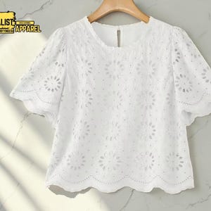 Può includere: Top bianco traforato con maniche corte e scollo tondo. Il top presenta un motivo floreale ed è appeso a una gruccia di legno. Il marchio "Minimalist Apparel" è visibile in alto a sinistra.