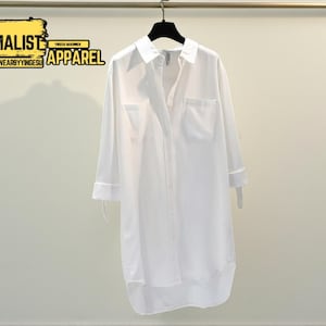 May include: A white shirt dress with long sleeves, a collared neckline, and two chest pockets, hanging on a black hanger. The dress is buttoned down the front. The text "MINIMALIST APPAREL" is visible in the upper left corner.
