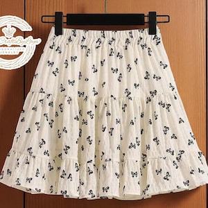 May include: A cream-colored skirt with a black butterfly pattern. The skirt has a tiered, ruffled design and an elastic waistband. The skirt is hanging on a black hanger against a wooden background.