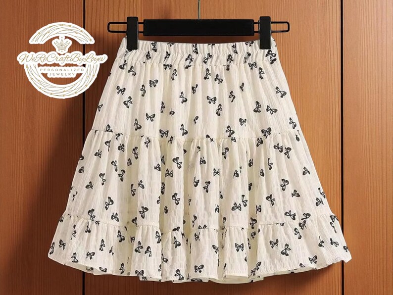May include: A cream-colored skirt with a black butterfly pattern. The skirt has a tiered, ruffled design and an elastic waistband. The skirt is hanging on a black hanger against a wooden background.