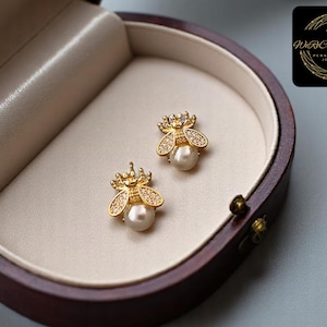May include: A pair of gold-colored bee-shaped stud earrings with pearl-like centers. The bees have crystal accents on their wings and crowns. The earrings are displayed in a brown jewelry box with a cream-colored interior.