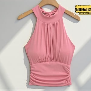 Könnte beinhalten: Ein hellrosa Neckholder-Top mit hohem Ausschnitt und geraffter Brustpartie. Das Top hängt an einem Holzbügel vor weißem Hintergrund. Der Text "MINIMALIST APPAREL" ist in der oberen rechten Ecke sichtbar.