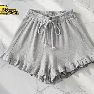 Può includere: Pantaloncini grigio chiaro con orlo a balze e vita con coulisse. I pantaloncini hanno un aspetto a maglia a nido d'ape strutturato. L'immagine include anche il testo "MINIMALIST APPAREL" in un logo rettangolare giallo e nero.