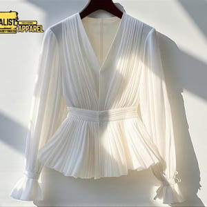 May include: A white, long-sleeved blouse with a V-neck and pleated bodice. The blouse features a peplum waist, button details, and flared cuffs. The image includes the text "MINIMALIST APPAREL".
