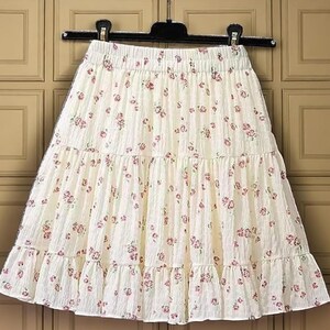 May include: Cream-colored tiered skirt with a floral pattern. The skirt is hanging on a black hanger. The fabric has a textured appearance, and the floral print features small, pink flowers scattered across the surface. The skirt has a ruffled hem.