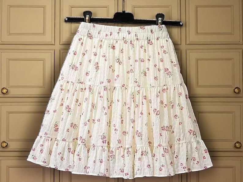 May include: Cream-colored tiered skirt with a floral pattern. The skirt is hanging on a black hanger. The fabric has a textured appearance, and the floral print features small, pink flowers scattered across the surface. The skirt has a ruffled hem.