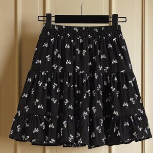 May include: Black tiered skirt with a white butterfly pattern. The skirt is hanging on a black hanger. The skirt has an elastic waistband and a ruffled hem. The skirt is made of a lightweight fabric.