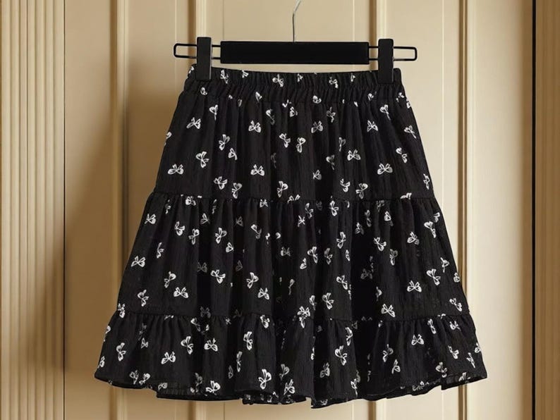 May include: Black tiered skirt with a white butterfly pattern. The skirt is hanging on a black hanger. The skirt has an elastic waistband and a ruffled hem. The skirt is made of a lightweight fabric.