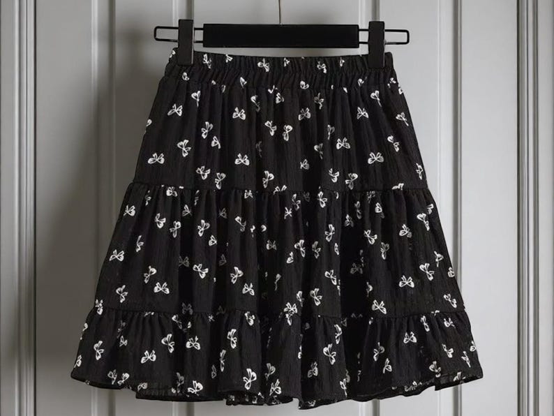 May include: Black tiered skirt with white butterfly pattern. The skirt is hanging on a black hanger. The skirt has a ruffled hemline and an elastic waistband.