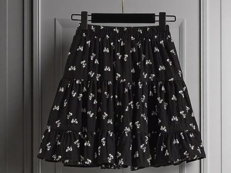 May include: Black tiered skirt with white butterfly pattern. The skirt is hanging on a black hanger. The skirt has an elastic waistband and a ruffled hem. The skirt is made of a lightweight fabric.