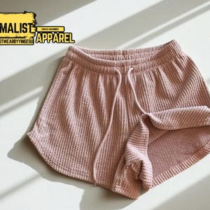May include: Pale pink waffle-knit shorts with a drawstring waist and rolled hem. The shorts are made of a textured fabric. The image includes the text "MINIMALIST APPAREL" and "MODESTWEARBYYINGESU".