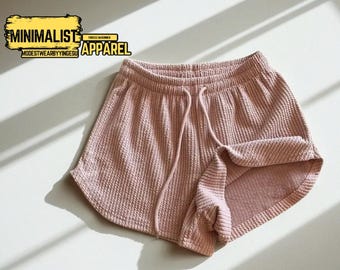 May include: Pink waffle-knit shorts with a drawstring waist. The shorts have a rolled hem and are made of a textured fabric. The image includes the text "MINIMALIST APPAREL" and "MODESTWEARBYYINGESU".