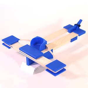 May include: A blue and wood toy catapult with a rubber band and a blue base. The catapult is designed to launch small objects.