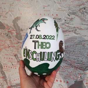 Pode incluir: Um ovo branco com ilustra&ccedil;&otilde;es de dinossauros e outros motivos em verde, azul e preto. O ovo tem o texto "27.08.2022 THEO EINSCHULUNG" escrito nele.