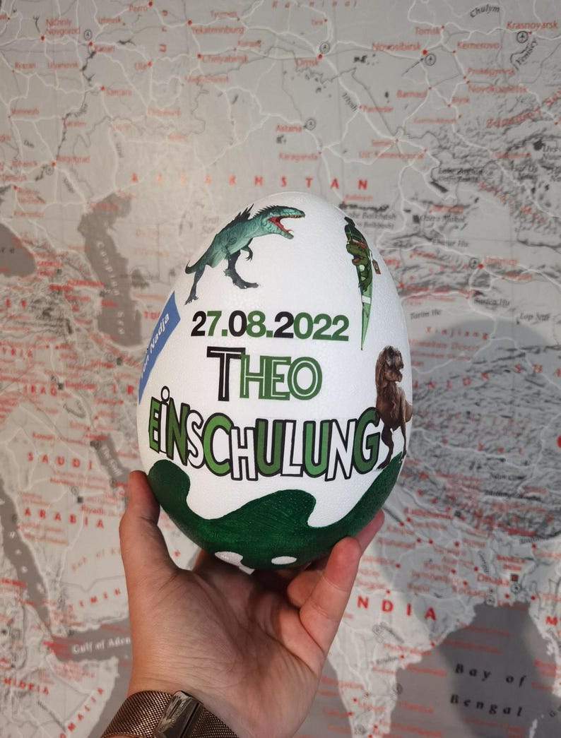 Pode incluir: Um ovo branco com ilustra&ccedil;&otilde;es de dinossauros e outros motivos em verde, azul e preto. O ovo tem o texto "27.08.2022 THEO EINSCHULUNG" escrito nele.