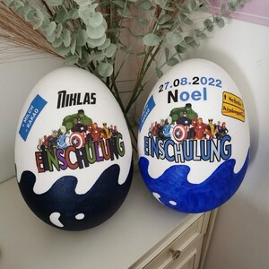 Pode incluir: Dois ovos brancos decorados com tinta azul e branca. Um ovo tem o nome "Niklas" e o texto "Einschulung" com um tema de super-her&oacute;i. O outro ovo tem a data "27.08.2022" e o nome "Noel" com o texto "Einschulung" e um tema de super-her&oacute;i.