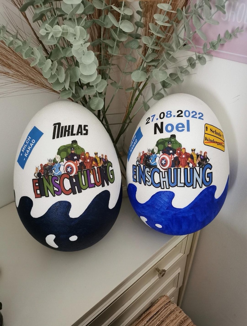 Pode incluir: Dois ovos brancos decorados com tinta azul e branca. Um ovo tem o nome "Niklas" e o texto "Einschulung" com um tema de super-her&oacute;i. O outro ovo tem a data "27.08.2022" e o nome "Noel" com o texto "Einschulung" e um tema de super-her&oacute;i.