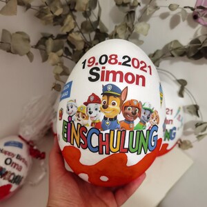Pode incluir: Um ovo de chocolate branco com um design da Patrulha Canina. O ovo &eacute; decorado com o texto "19.08.2021 Simon Einschulung".