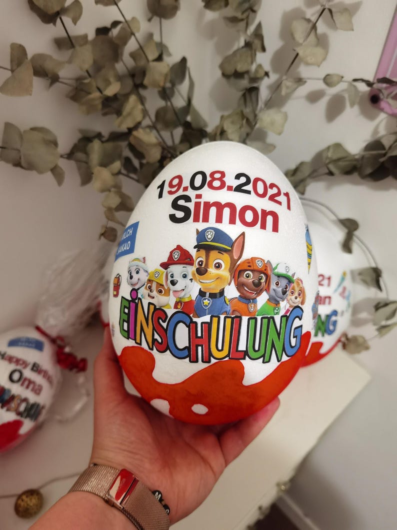 Pode incluir: Um ovo de chocolate branco com um design da Patrulha Canina. O ovo &eacute; decorado com o texto "19.08.2021 Simon Einschulung".
