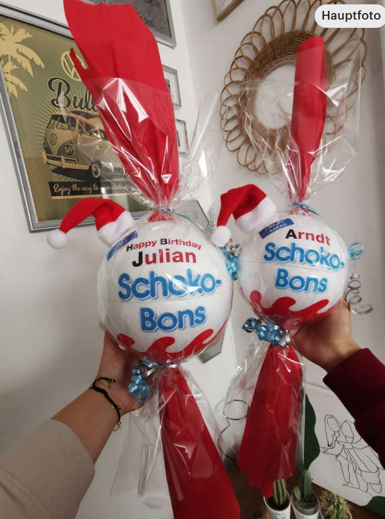 May include: Two white and red Schoko-Bons gifts, each wrapped in clear cellophane and red paper. Each gift has a small Santa hat and a blue ribbon. One says "Happy Birthday Julian", the other says "Arndt".