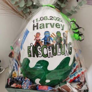Pode incluir: Uma sacola de presente branca com um design verde e branco com o texto "11.08.2022 Harvey Einschulung". A sacola est&aacute; cheia de v&aacute;rias barras de chocolate e doces.