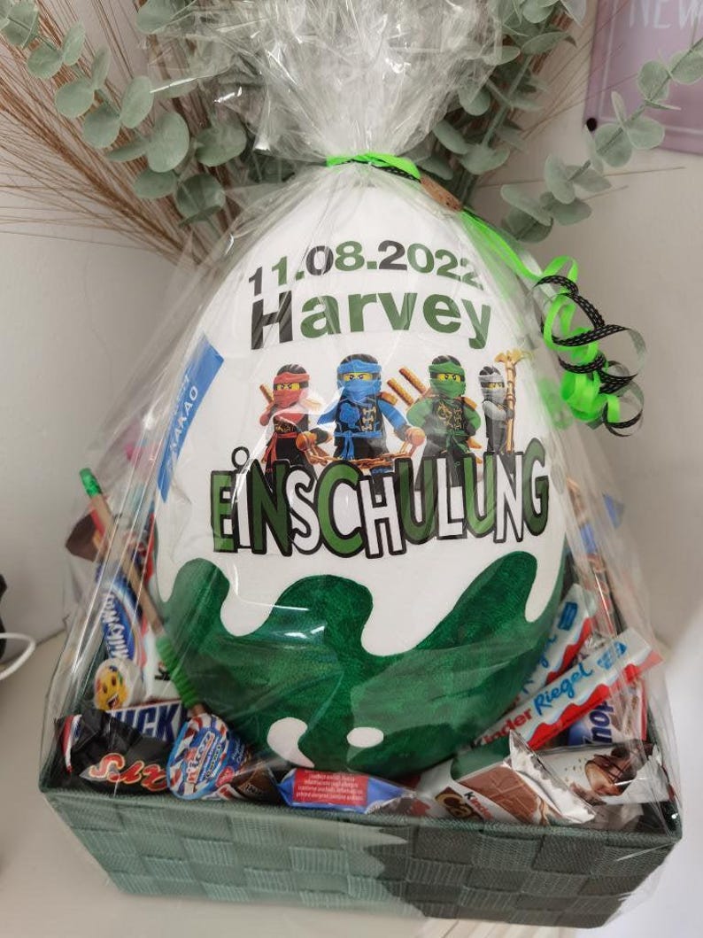 Pode incluir: Uma sacola de presente branca com um design verde e branco com o texto "11.08.2022 Harvey Einschulung". A sacola est&aacute; cheia de v&aacute;rias barras de chocolate e doces.