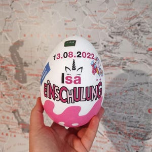 Pode incluir: Objeto branco em forma de ovo com bolinhas rosa e brancas. O ovo tem um adesivo de unic&oacute;rnio e o texto "13.08.2022 Isa Einschulung" nele.