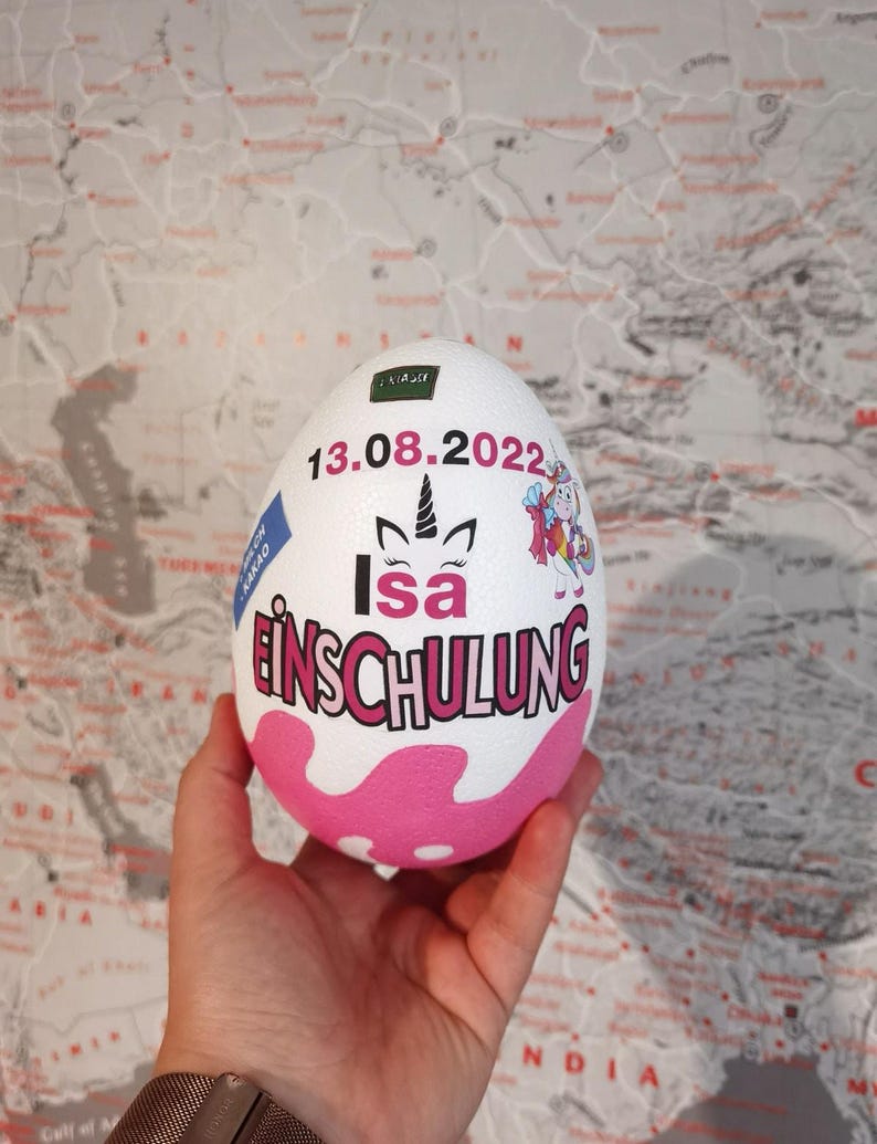 Pode incluir: Objeto branco em forma de ovo com bolinhas rosa e brancas. O ovo tem um adesivo de unic&oacute;rnio e o texto "13.08.2022 Isa Einschulung" nele.
