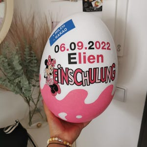 Pode incluir: Uma decora&ccedil;&atilde;o em forma de ovo rosa e branco com uma imagem de desenho animado da Minnie Mouse. O ovo tem o texto "MILCH - KAKAO" e "06.09.2022 Elien EINSCHULUNG".
