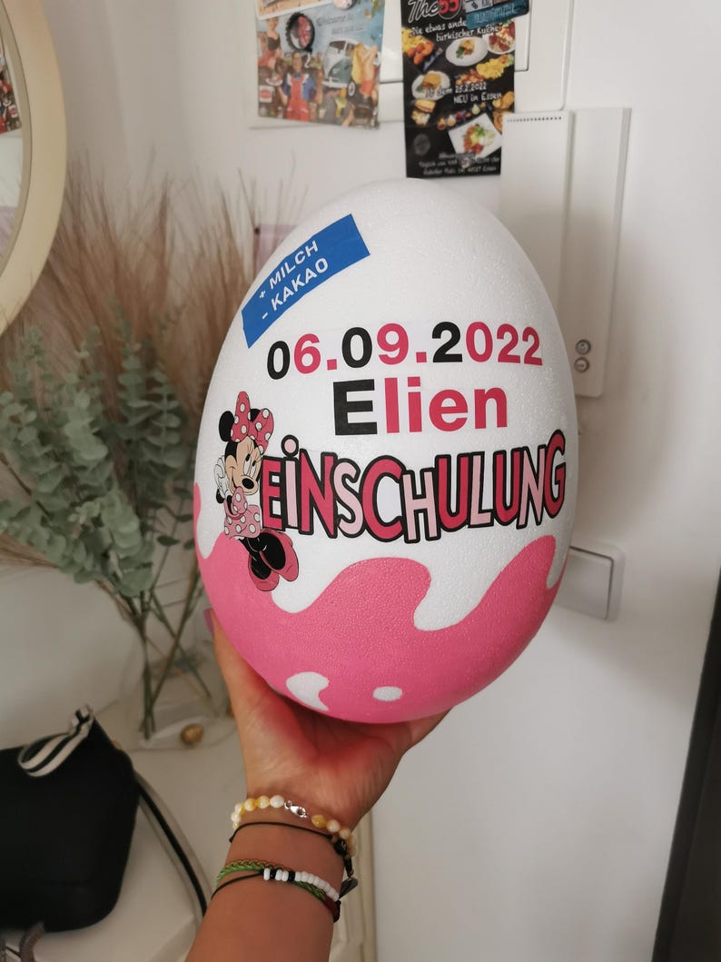 Pode incluir: Uma decora&ccedil;&atilde;o em forma de ovo rosa e branco com uma imagem de desenho animado da Minnie Mouse. O ovo tem o texto "MILCH - KAKAO" e "06.09.2022 Elien EINSCHULUNG".