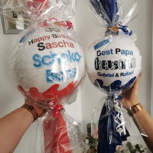 May include: Two white and red Kinder Surprise egg gifts, wrapped in clear cellophane. One egg has "Happy Birthday Sascha" printed on it, the other says "Best Papa". Both are tied with ribbons and bows.