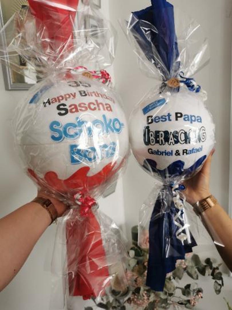 May include: Two white and red Kinder Surprise egg gifts, wrapped in clear cellophane. One egg has "Happy Birthday Sascha" printed on it, the other says "Best Papa". Both are tied with ribbons and bows.