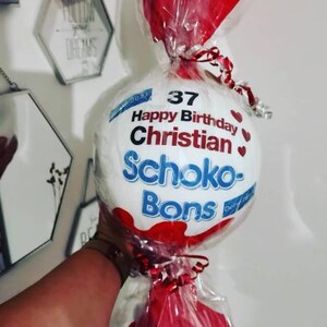 May include: A white, round candy package with the words "Happy Birthday Christian" and "Schoko-Bons" printed on it. The package is wrapped in clear cellophane and tied with red ribbon. Red decorative elements are also included.