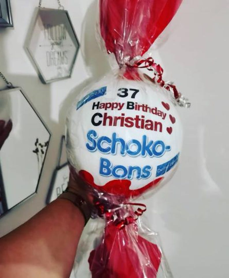 May include: A white, round candy package with the words "Happy Birthday Christian" and "Schoko-Bons" printed on it. The package is wrapped in clear cellophane and tied with red ribbon. Red decorative elements are also included.