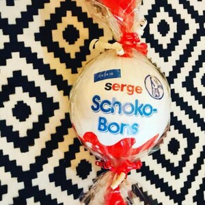 May include: A large, white, round candy wrapped in clear cellophane and tied with red ribbon. The candy has the words "serge Schoko-Bons" in blue and red. A small blue sticker with the date "09.09.18" is visible.