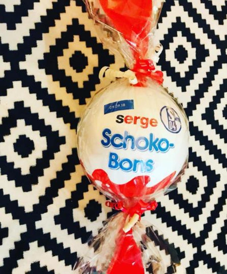 May include: A large, white, round candy wrapped in clear cellophane and tied with red ribbon. The candy has the words "serge Schoko-Bons" in blue and red. A small blue sticker with the date "09.09.18" is visible.