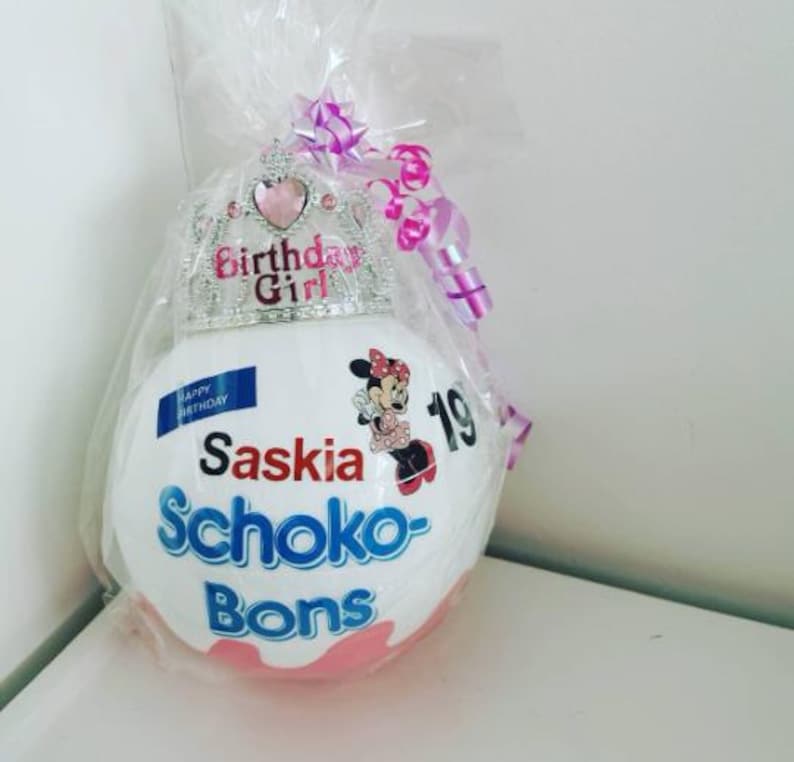 May include: A white, egg-shaped gift wrapped in clear cellophane, adorned with a silver tiara and pink bow. The gift features the text "Birthday Girl", "Saskia", "Schoko-Bons", and a Minnie Mouse graphic. A sticker indicates the number "19".