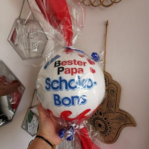 May include: A white and blue ball-shaped gift wrapped in clear cellophane and red fabric. The text "Bester Papa Schoko-Bons" is printed on the ball. Blue and white ribbons are tied around the base of the gift.