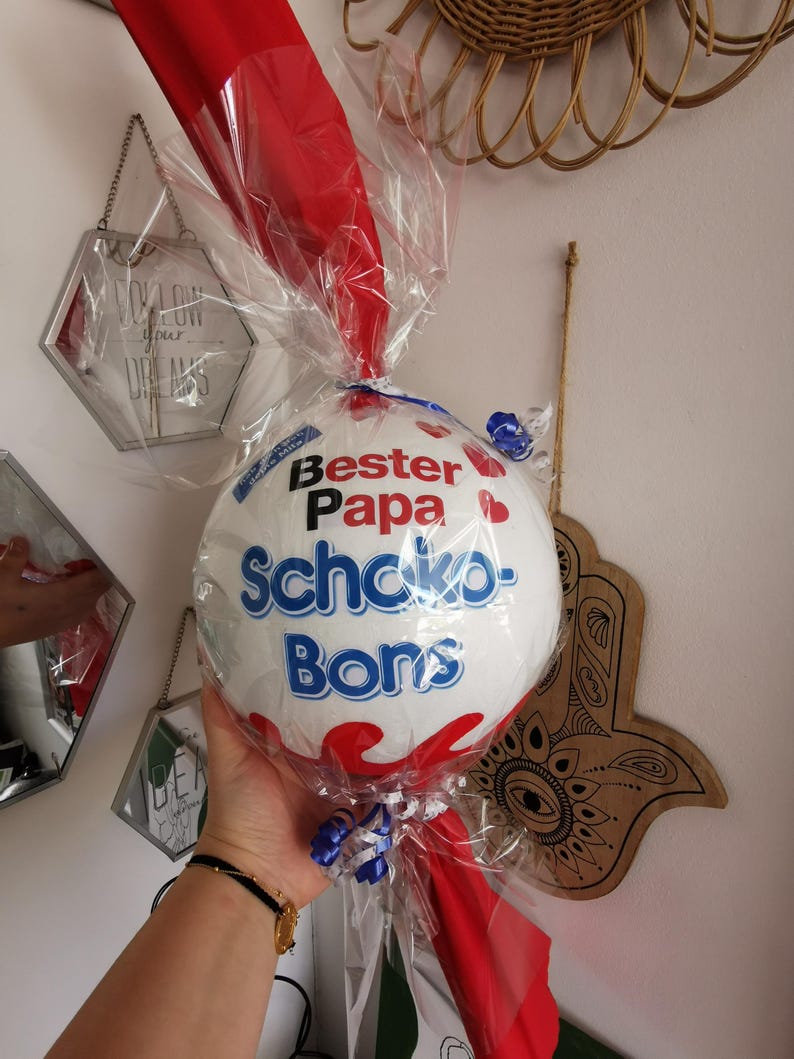 May include: A white and blue ball-shaped gift wrapped in clear cellophane and red fabric. The text "Bester Papa Schoko-Bons" is printed on the ball. Blue and white ribbons are tied around the base of the gift.