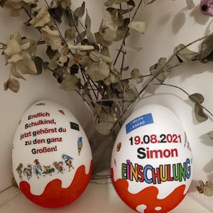 Pode incluir: Dois ovos de chocolate branco decorados com tinta vermelha e branca. Um ovo tem o texto "Endlich Schulkind, jetzt geh&ouml;rst du zu den ganz Gro&szlig;en!" e o outro ovo tem o texto "MILCH KAKAO 19.08.2021 Simon EINSCHULUNG".