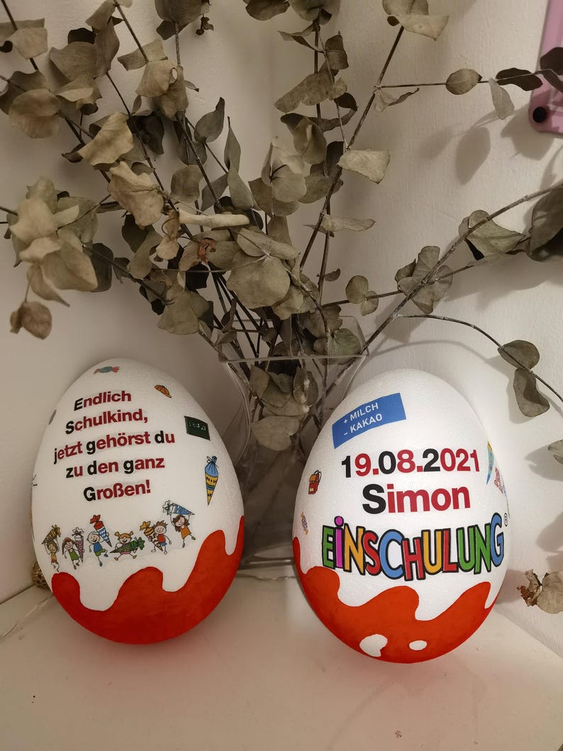 Pode incluir: Dois ovos de chocolate branco decorados com tinta vermelha e branca. Um ovo tem o texto "Endlich Schulkind, jetzt geh&ouml;rst du zu den ganz Gro&szlig;en!" e o outro ovo tem o texto "MILCH KAKAO 19.08.2021 Simon EINSCHULUNG".