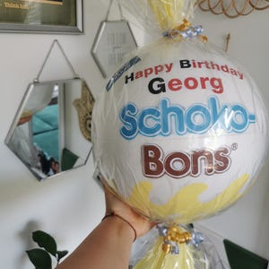 May include: A white, round gift wrapped in clear cellophane, featuring the text "Happy Birthday Georg" and the brand name "Schoko-Bons". The gift is decorated with yellow and blue ribbons.