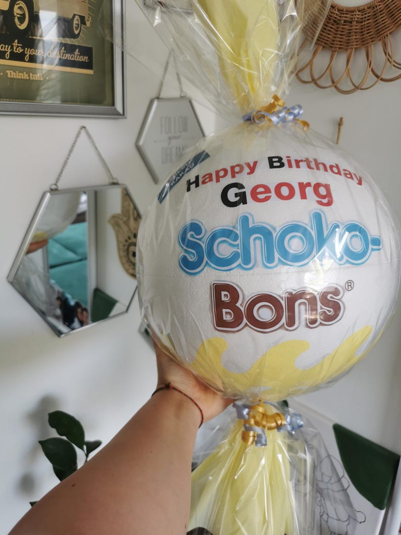 May include: A white, round gift wrapped in clear cellophane, featuring the text "Happy Birthday Georg" and the brand name "Schoko-Bons". The gift is decorated with yellow and blue ribbons.