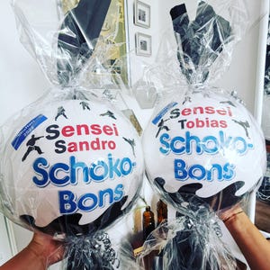 May include: Two white, round gift baskets with the words "Sensei Sandro Schoko-Bons" and "Sensei Tobias Schoko-Bons" in blue and red lettering. Each basket is wrapped in clear cellophane and tied with a black bow.
