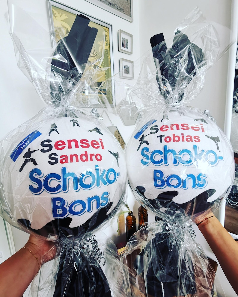 May include: Two white, round gift baskets with the words "Sensei Sandro Schoko-Bons" and "Sensei Tobias Schoko-Bons" in blue and red lettering. Each basket is wrapped in clear cellophane and tied with a black bow.