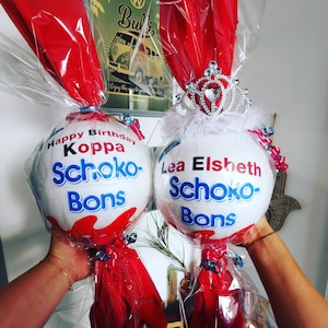 May include: Two white, egg-shaped gifts wrapped in clear cellophane and red tissue paper. One is topped with a silver tiara and the other has text that reads "Happy Birthday Koppa Schoko-Bons". Both are held up by hands.