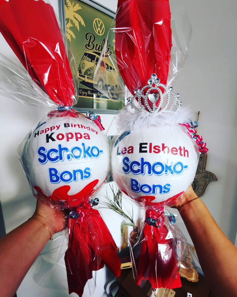 May include: Two white, egg-shaped gifts wrapped in clear cellophane and red tissue paper. One is topped with a silver tiara and the other has text that reads "Happy Birthday Koppa Schoko-Bons". Both are held up by hands.