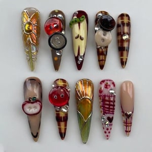 May include: A collection of ten long, almond-shaped artificial nails. Each nail features unique designs, including floral patterns, fruit-themed embellishments, and plaid accents. Some nails are adorned with rhinestones and buttons, showcasing a variety of colors and textures.