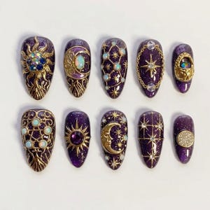 May include: A set of ten purple and gold celestial-themed artificial nails. Each nail features intricate designs, including suns, moons, stars, and opalescent accents. The nails are oval-shaped and have a glossy finish.